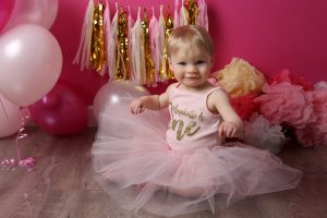 Little girl in pink tutu personalised outfit for cake smash photoshoot in east grinstead with pink backdrop and props