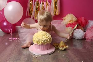 Little girl eating pink and cream cake with wooden spoon with pink backdrop and balloons and pom poms at photoshoot in West Sussex
