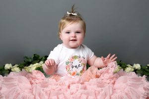 little girl celebrating first birthday in pink tutu and number one babygrow with grey backdrop and floral prop at photoshoot in west sussex
