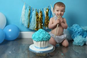 little boy holding wooden spoon at cakesmash photography session with blue backdrop and blue baloons and props