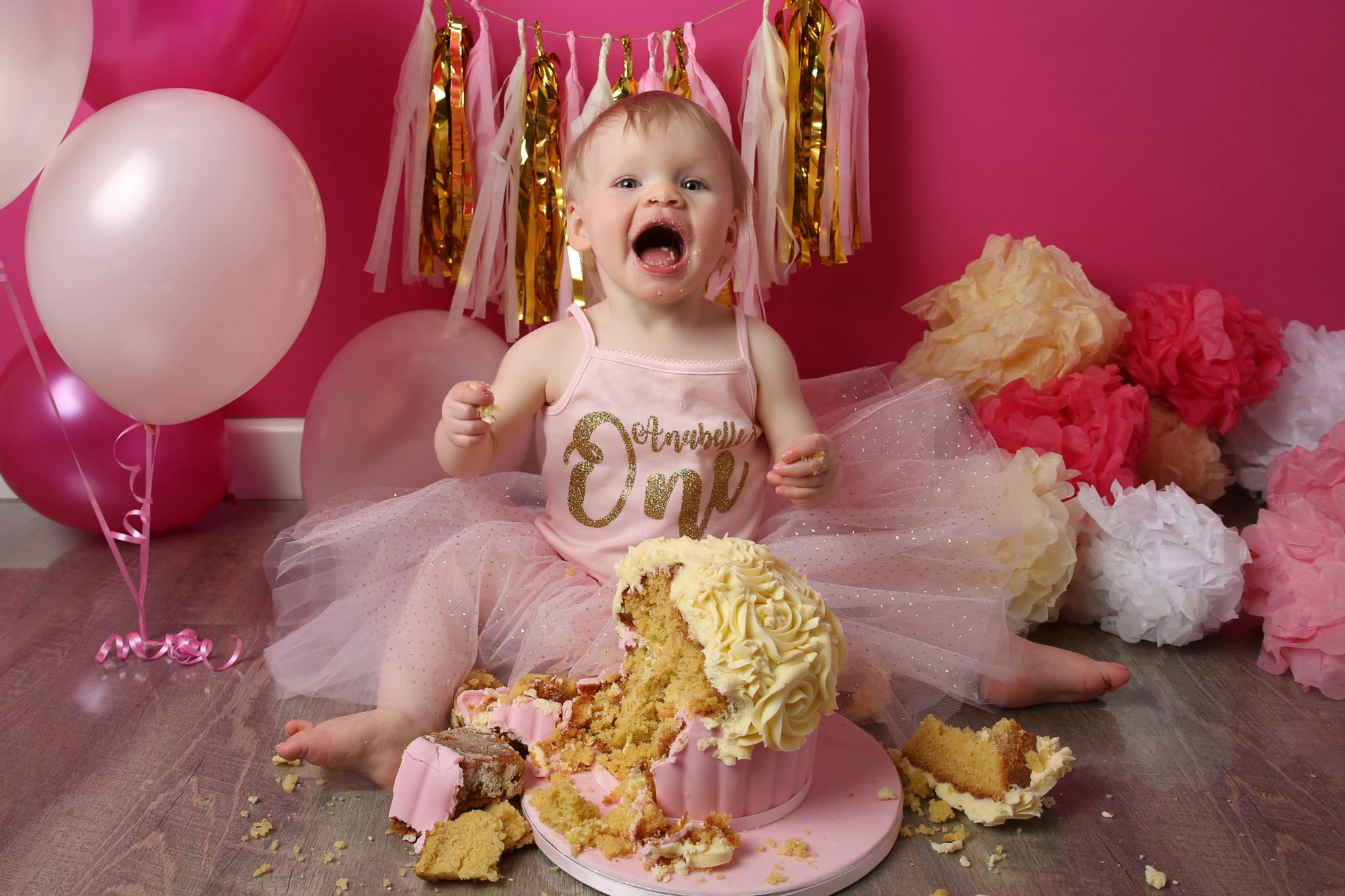 little girl excited about eating cake at photography session in west sussex with pink backdrop and balloons and pom poms