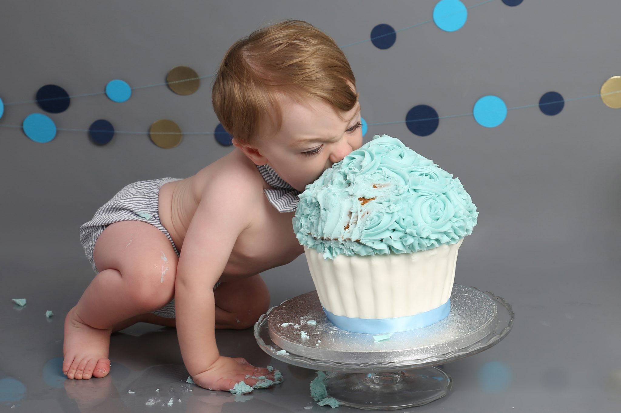 baby boy eating blue and white cake head first blue banner and grey backdrop at cake smash photoshoot west sussex