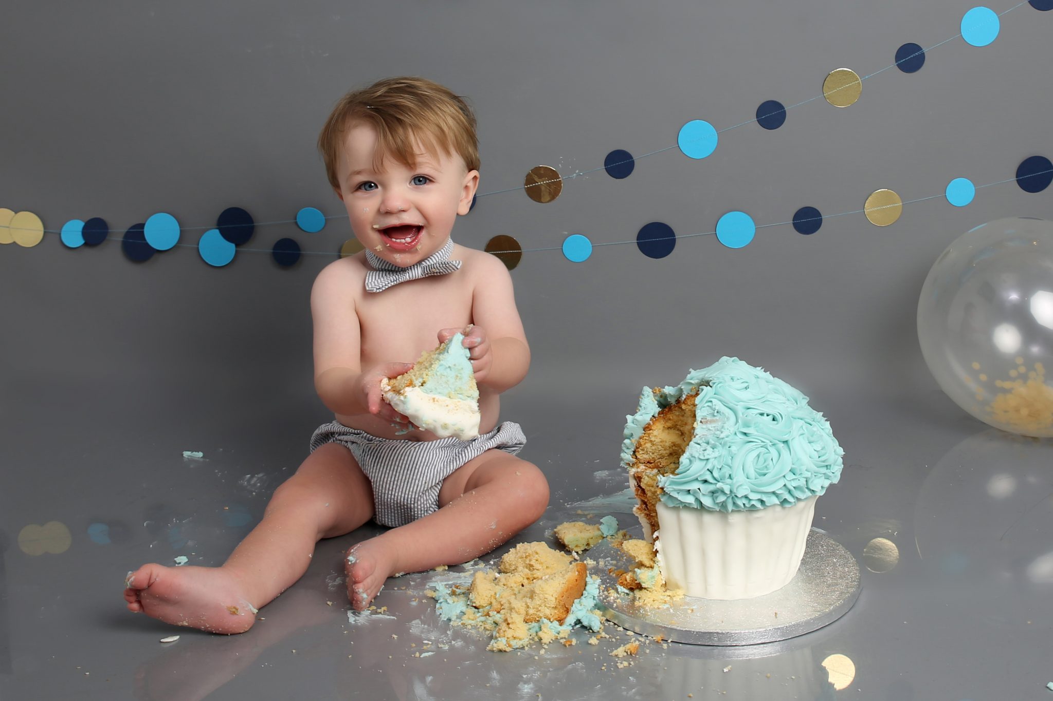 little boy holding blue and white cake at cakesmash photoshoot in east grinstead with grey backdrop and blue and gold banner