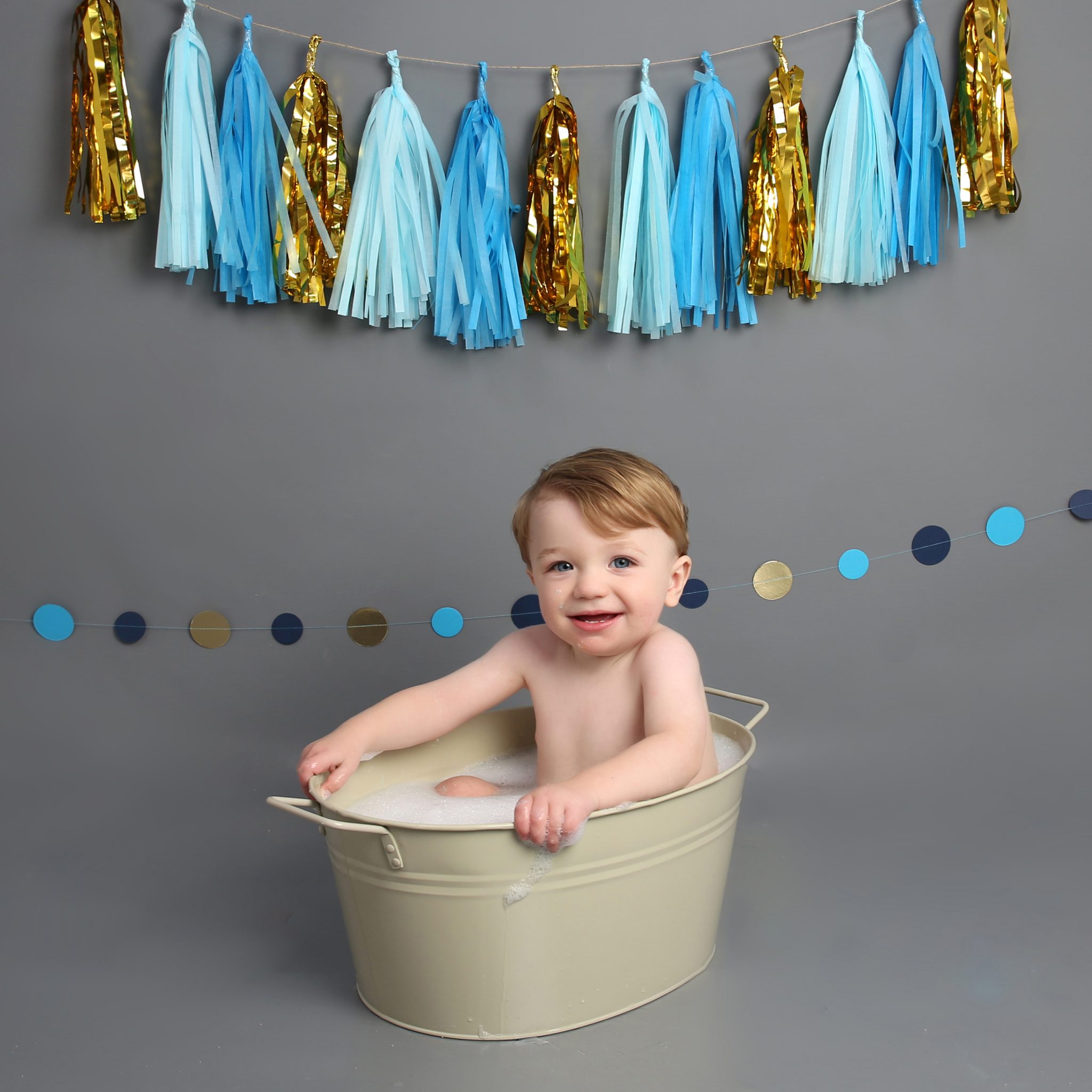 Little boy smiling in cream bath tub with blue and gold banner at cake smash photoshoot West Sussex