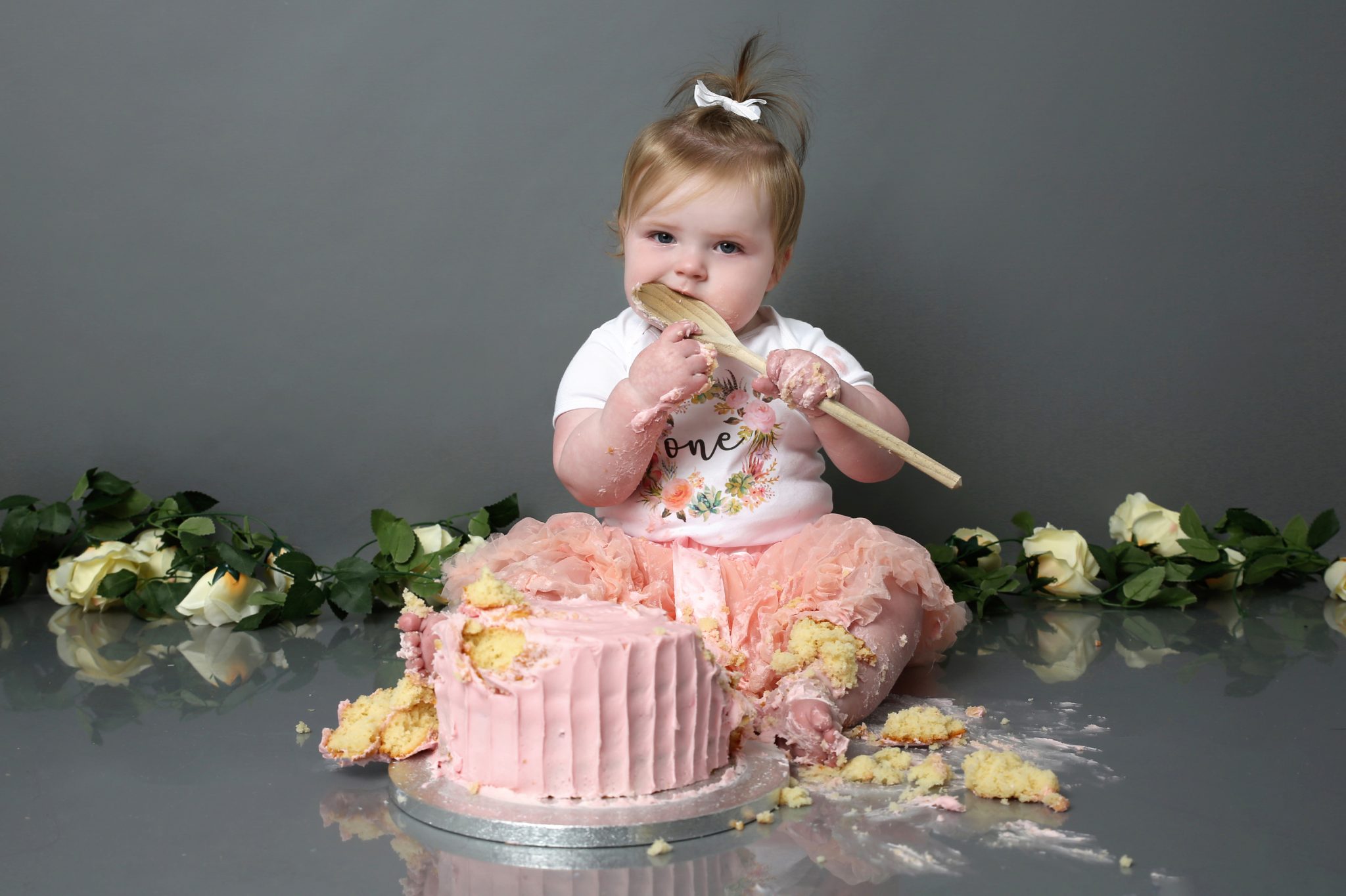 Little girl eating cake of wooden spoon sitting with pink cake with floral prop at cake smash photoshoot West Sussex