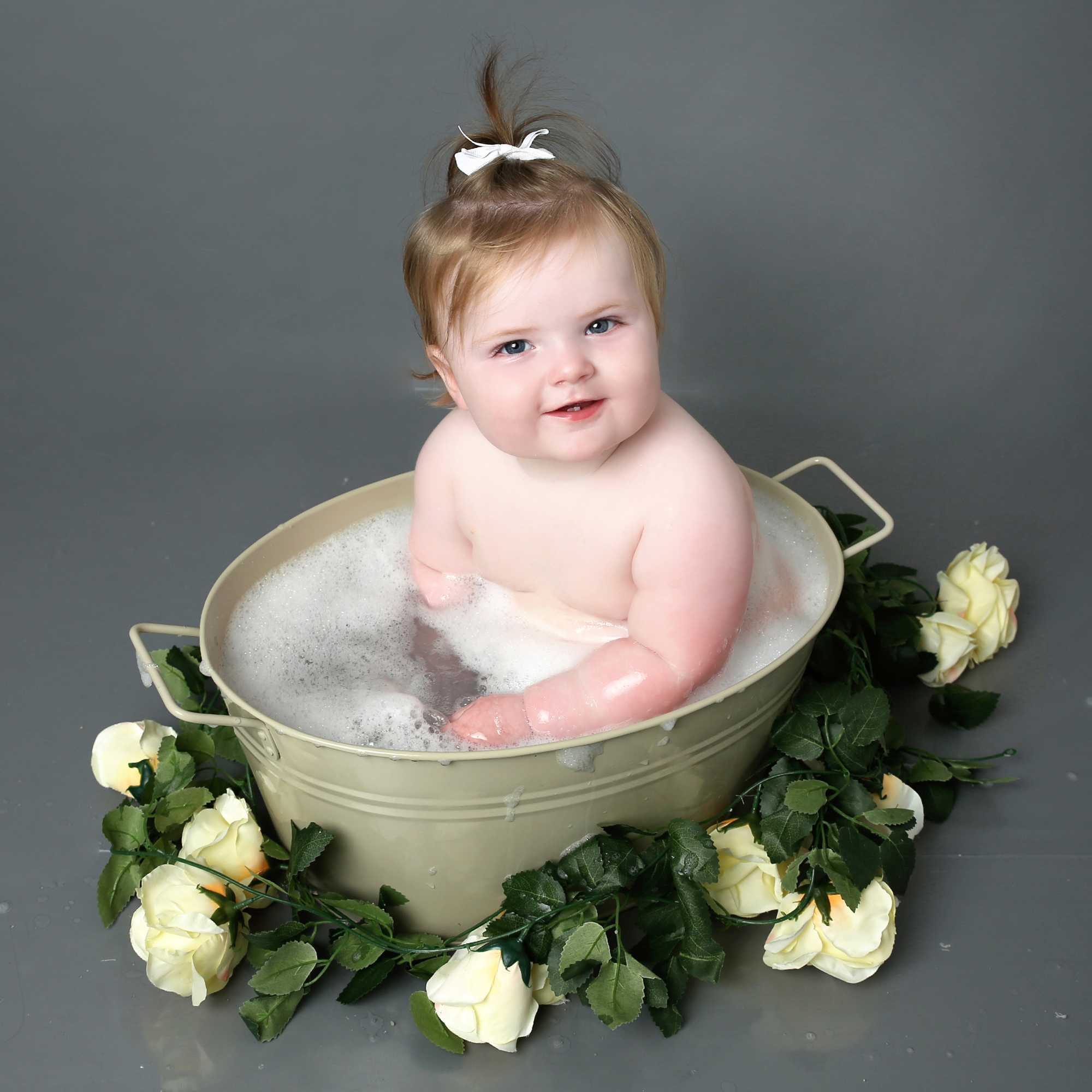 Little girl sitting in bath tub smiling at camera with grey backdrop and floral prop at cake smash photoshoot West Sussex