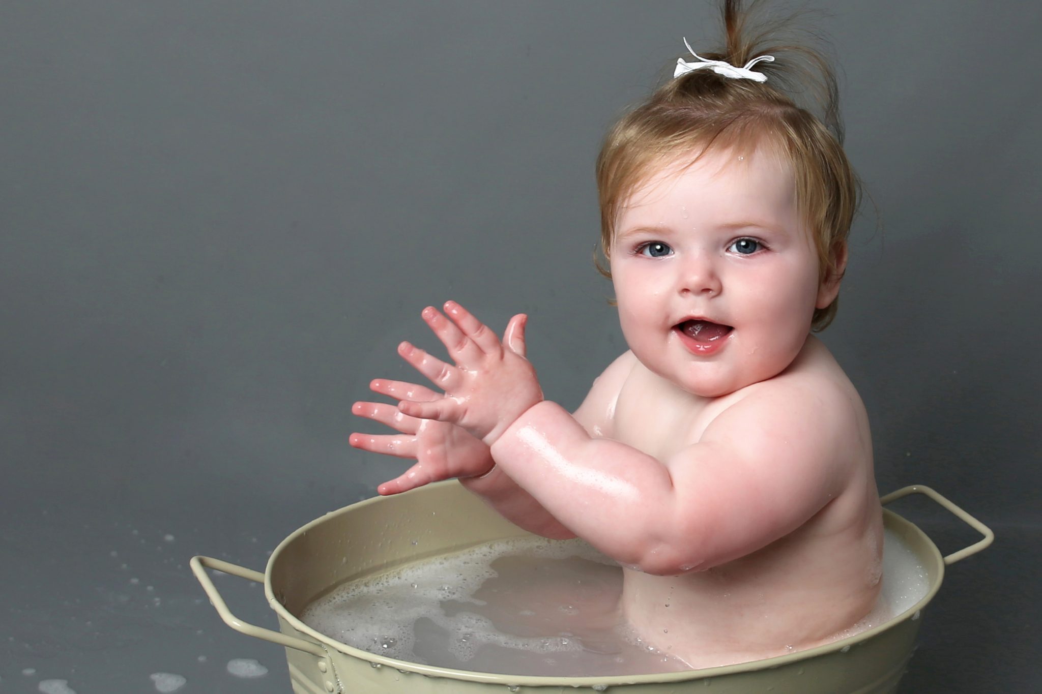 happy little girl in bath tub clapping hands with grey backdrop at cakesmash photoshoot in east grinstead