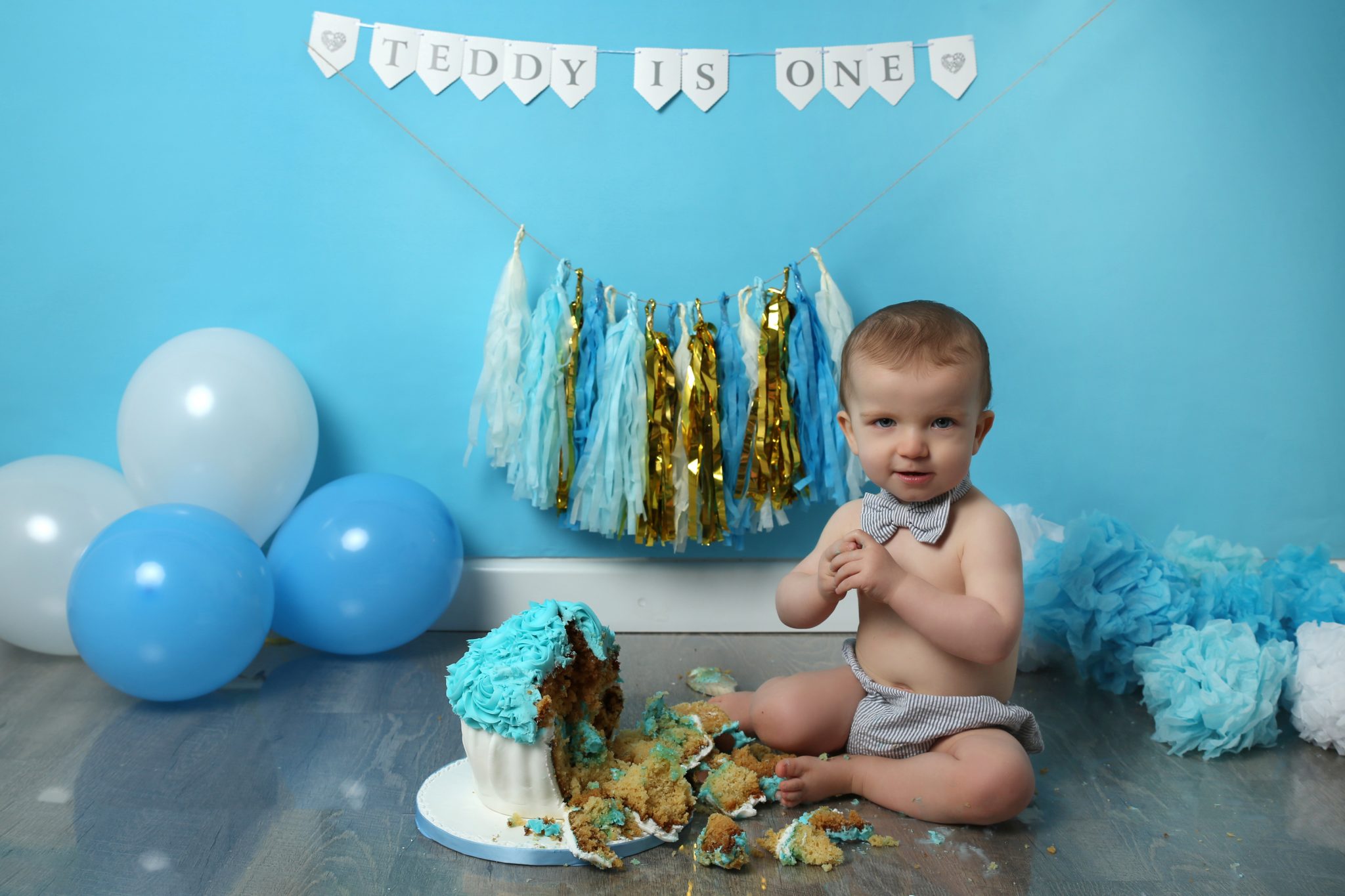 little boy at cakesmash photoshoot in west sussex with smashed blue and white cake and blue balloons and props